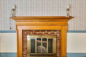 A wooden fireplace mantel with a brick surround and a brass-framed glass fireplace screen. Two glass candle holders with white candles are placed at each end of the mantel. The wall has patterned wallpaper.