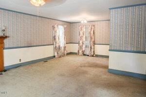 Empty living room with patterned wallpaper, cream carpet, blue trim, and floral curtains on two windows and a door. A wooden fireplace mantel is partially visible on the left side of the image. Ceiling lights are on.