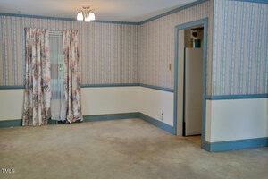 A room with patterned wallpaper, blue trim, and floral curtains over a window. The carpet is beige and stained. A doorway on the right leads to another space. There is a ceiling light fixture above.