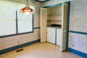 A small laundry area with a washer and dryer in a closet, located in a room with patterned wallpaper, blue trim, a window with sheer curtains, and a stained glass ceiling light.