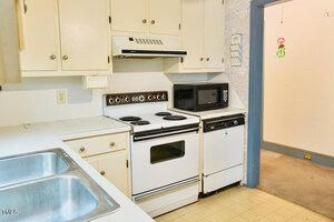A small kitchen with a white electric stove, oven, and range hood, beige cabinets, a black microwave, and a double stainless steel sink. The floor is yellowed tile, and an adjoining room is visible through a doorway.