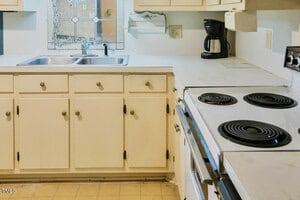 A kitchen with white cabinets, a double sink under a window, a black coffee maker, an electric stove with coil burners, and a beige countertop. The floor has a light-colored square tile pattern.