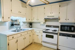 A small kitchen with cream-colored cabinets, a double sink, electric stove, microwave, dishwasher, coffee maker, and a ceiling fan. The countertop is white and the floor is light-colored linoleum.