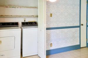 A laundry area with a white dryer and washing machine beneath two bare shelves, next to a wall with blue trim and patterned wallpaper. The space has linoleum flooring and visible electrical outlets.