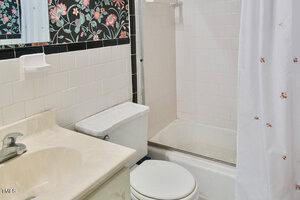 A bathroom with a beige sink and countertop, white toilet, and a white tiled shower with a curtain. The wall above the sink has floral wallpaper and a mirror. The shower has a built-in soap holder.