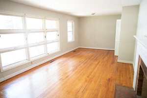 A bright, empty living room with large windows on one wall, hardwood floors, beige walls, a white fireplace, and a small ceiling light fixture.