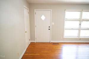 A white front door with a diamond-shaped window is set in a light gray wall. To the left of the door is a small closed closet. To the right, there are four rectangular windows. The floor is hardwood.