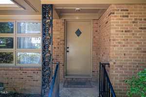 A beige door with a diamond-shaped window is centered in a brick entryway with black metal railings and a decorative black metal post. A doormat is in front of the door, and windows are visible to the left.