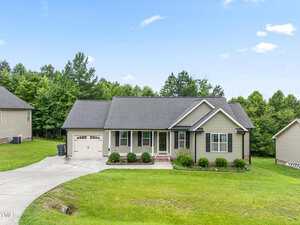 A single-story house at 421 Angus Place in Stem features gray siding, a dark roof, an attached single-car garage, front porch with white columns, and a well-kept lawn on a sloped driveway with trees and other homes in the background.