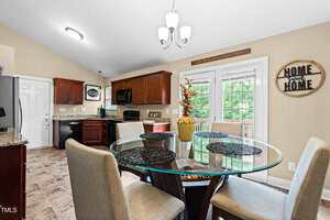 A kitchen with brown cabinets, granite countertops, and stainless steel appliances. A round glass dining table with four chairs is in the foreground, and a door leads to a balcony. Wall decor includes home-themed signs.