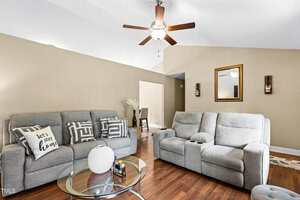 A living room with gray sofas, a glass coffee table with a white vase, wooden floors, beige walls, a ceiling fan, and wall decor including a mirror and two sconces. A pillow reads "Let's stay home.