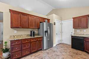 A kitchen with brown wood cabinets, granite countertops, a stainless steel refrigerator, a black dishwasher, a coffee maker, tiled flooring, and a potted plant in the corner.