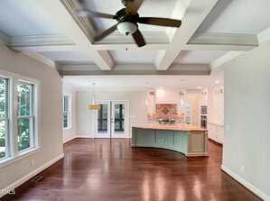 A spacious open-plan room with wood floors, a coffered ceiling with a ceiling fan, large windows, a kitchen with an island and pendant lights, and a dining area near glass doors leading outside.
