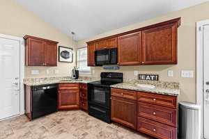 A kitchen with brown wooden cabinets, granite countertops, black appliances including a stove and dishwasher, beige walls, tile flooring, and a window above the sink. A decorative sign reads "EAT.