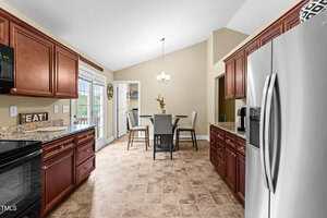 A kitchen with brown cabinets, granite countertops, and stainless steel appliances opens into a dining area with a round table and four chairs. A chandelier hangs from the ceiling, and French doors lead outside.