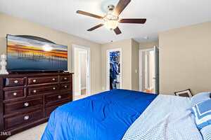A bedroom with beige walls, a ceiling fan, a large dresser with a flat-screen TV, a bed with blue and white bedding, and open doors revealing a closet and a hallway.