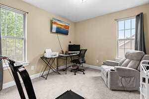 A home office with a desk holding a computer, printer, and office supplies. There is a black office chair, a gray recliner, two windows with curtains, beige walls, and a wall-mounted TV displaying a sunset.
