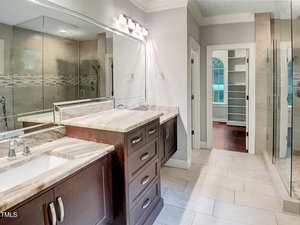 Modern bathroom with dual sinks, dark wood cabinets, marble countertops, glass-enclosed shower, light tile floor, large mirror with overhead lighting, and an open doorway leading to a walk-in closet with shelves.