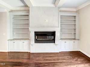 A white fireplace with a dark glass insert is centered between two built-in shelving units with cabinets below. The room has light walls, crown molding, and dark hardwood floors.