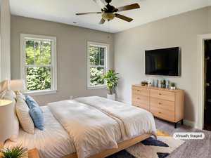 A modern bedroom with a large bed, wooden dresser, wall-mounted TV, two windows, a plant, and a ceiling fan. The room has light-colored walls and is bright with natural light. A "Virtually Staged" label appears in the corner.