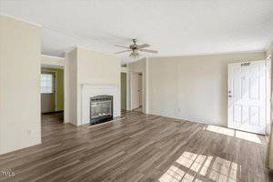 A bright, empty living room with light wood flooring, beige walls, a white ceiling fan, a white fireplace, and a white front door. Sunlight streams through windows onto the floor.