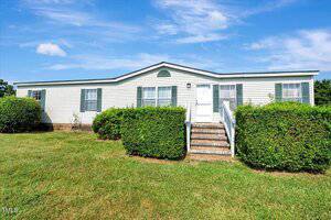 Single-story manufactured home with white siding, green shutters, and a front entrance accessed by steps and a metal railing. Bushes line the walkway, and the house sits on a grassy lawn under a blue sky.