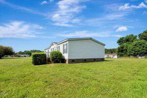 A single-story white manufactured home with multiple windows sits on a large grassy lot under a blue sky with scattered clouds. Bushes are planted along the front of the house. Trees and other homes are visible in the background.