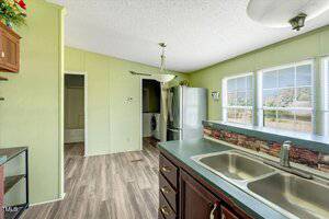 A kitchen with green walls, wood cabinets, a double sink, stainless steel refrigerator, and a partial view of a laundry area and bathroom. Large windows let in natural light. Wood-look flooring runs throughout.