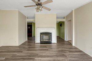 A room with wood-look flooring, beige walls, a white fireplace at the center, a ceiling fan, and open walkways leading to adjoining rooms.