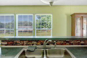 A stainless steel kitchen sink with a faucet and dishcloth sits beneath a green countertop. Behind the sink is a partial stone backsplash, three large windows with blinds, and a wooden cabinet to the right. Trees are visible outside.