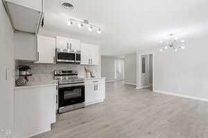A modern, open-concept kitchen and living area with white cabinets, stainless steel appliances, light wood flooring, neutral walls, and contemporary lighting fixtures. The space appears clean and bright.