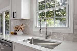A modern kitchen sink with a stainless steel faucet beneath a window. The countertop is white with a vase of flowers and a lemon at the left. Light gray tile backsplash and white cabinetry are visible.