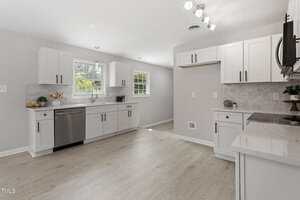 A modern kitchen with white cabinets, stainless steel appliances, light wood flooring, white countertops, and two windows letting in natural light. Decor includes a fruit bowl and a vase with pink flowers.