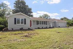 Charming single-story white brick house at 946 Jacksontown Road in Henderson, featuring a brown shingle roof, black shutters, red front door, attached two-car garage, and a spacious front lawn under blue skies with scattered clouds.