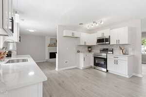 A modern kitchen features white cabinets, stainless steel appliances, light wood flooring, a white quartz countertop, and a partial view of a living area with a fireplace in the background.