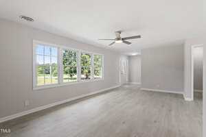 Spacious living room with light wood flooring, large windows showing a green outdoor view, white walls, and a ceiling fan with light. A doorway and main entrance are visible in the background.