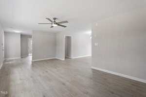 A spacious, empty living room with light wood flooring, beige walls, a ceiling fan with lights, and open doorways leading to other rooms in a modern home. Natural light enters from windows in the background.