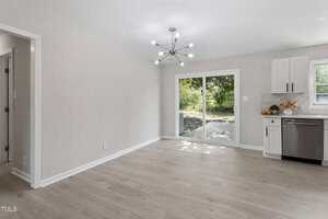A bright, empty dining area with light wood floors, a modern chandelier, and sliding glass doors leading to an outdoor deck. The kitchen with white cabinets and stainless steel appliances is partially visible on the right.