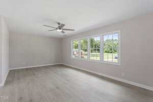 A large, empty room with light-colored wood flooring, white walls, a ceiling fan with lights, and three large windows letting in natural light. Trees and a parked vehicle are visible outside.