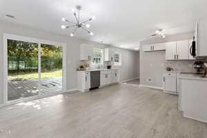 Bright, modern kitchen with white cabinets, light wood flooring, stainless steel appliances, and a sliding glass door opening to a backyard deck. Large windows let in natural light. Contemporary light fixtures are on the ceiling.