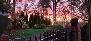 A backyard garden with green shrubs, black metal fencing, and brick pillars is shown at sunset. Trees and a pink, orange, and purple sky appear in the background. Two large planters with plants are visible in the foreground.