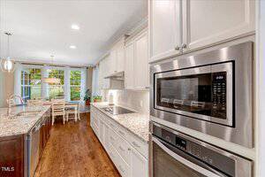 Modern kitchen with stainless steel appliances, white cabinets, granite countertops, and wood floors. A dining table with four chairs is placed by large windows overlooking a green outdoor area.