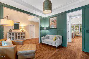 A living area with green paneled walls, wood floors, a gray loveseat with pillows, a tan armchair with ottoman, floor lamp, hanging light fixture, vintage poster, and view into a kitchen with shelves.