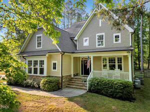 A two-story suburban house in Franklinton with beige siding, gray shingles, a gabled roof, front porch with white railings, stone foundation, and an American flag. 3650 Brooks Bend is surrounded by trees and greenery.