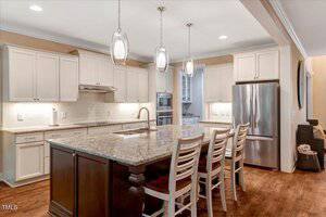 A modern kitchen with white cabinets, stainless steel appliances, granite countertops, a central island with a sink, and four wooden bar stools. Three pendant lights hang above the island, and the floor is hardwood.