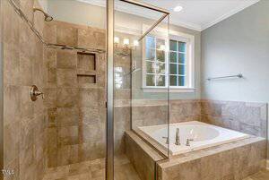 Bathroom with a glass-enclosed shower featuring beige tiles, a built-in shelf, and a bathtub with chrome fixtures next to a window. A towel rack is mounted on the wall.