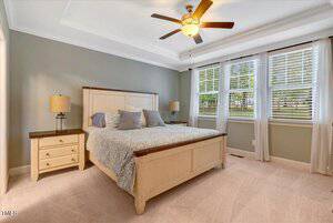 A bedroom with a wooden bed frame, light bedding, two nightstands with lamps, beige carpet, gray walls, a ceiling fan with a light, and three large windows with white blinds and curtains letting in natural light.