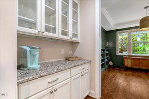 A granite countertop with white cabinets above and below, a light blue Keurig coffee maker, and a tray with two glasses. The area leads into a room with wood floors, shelves, and windows.