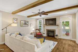 A living room with beige walls, wood floor, exposed ceiling beams, a white sectional sofa, a fireplace with a TV above it, a floor lamp, two windows, and a glass door leading outside.