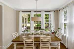A dining room with a wooden table and six chairs, a large round pendant light above, and several plants on the windowsill. Large windows let in natural light and provide a view of trees and greenery outside.